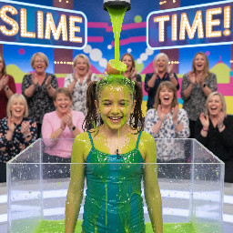 Tween girl with shoulder length brown hair in two high pigtails. She has a timid laugh of embarressment. She is wearing a blue spagetti top. A broken egg is on the top of her head, dripping yolk and bits of eggshell.