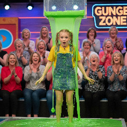 Youthful looking girl with blond hair in two high pigtail. Wearing short skirtall over yellow t shirt. Seems surprised. Hands besides her body.