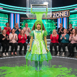 A young girl with brown hair dressed like alice in wonderland from the books version, looking slightly disgusted, embarrassed and surprised from the gunge.