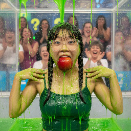 Asian young teen girl with short chin-length hair in two thick short braids ending at ear level, wearing glasses. She is wearing a dark green spagetti strap top, and holds an intact large apple in her wide open mouth. Her hands are on her shoulders. Close up.