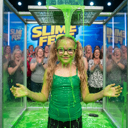 Tween with long sandy-blond wavy hair in high pigtails. Wearing tight green spagetti strap shirt. Has glasses with rectangular small frames. Playful shy smile with mouth partly open. Hands away from head. Photorealistic medium shot.