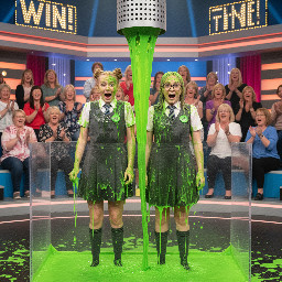 Two white teen girls. One has blond hair in two high tiny tight hair buns. The other has neck-length light ash brown hair and glasses. Both are wearing old-timey grey school uniform gymslips and have their mouths open in surprise.