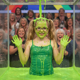 Tween with long sandy-blond wavy hair in high pigtails. Wearing tight green spagetti strap shirt. Has glasses with rectangular small frames. Playful shy smile with mouth partly open. Hands away from head. Photorealistic medium shot.