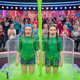 Two tween girls. One with medium length strawberry-blond hair covered with an elastic blue bandana. The other white with pale skin, large blue eyes and dark brown hair in two thick pigtail braids. Both wearing green tops. Both wearing glasses with thin small frames. Mouths open in playful surprise. Photorealistic.