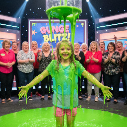 10 year old girl with round face, dirty blond hair and pale skin. Wearing a light blue t shirt and pink shorts. Is short and mostly skinny. Is smiling widely with hands to her sides at shoulder level, palms facing down and dripping slime.