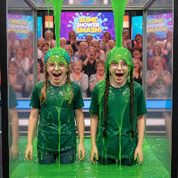 Two tween girls. One with medium length strawberry-blond hair covered with an elastic blue bandana. The other white with pale skin, large blue eyes and dark brown hair in two thick pigtail braids. Both wearing green tops. Both wearing glasses with thin small frames. Mouths open in playful surprise. Photorealistic.